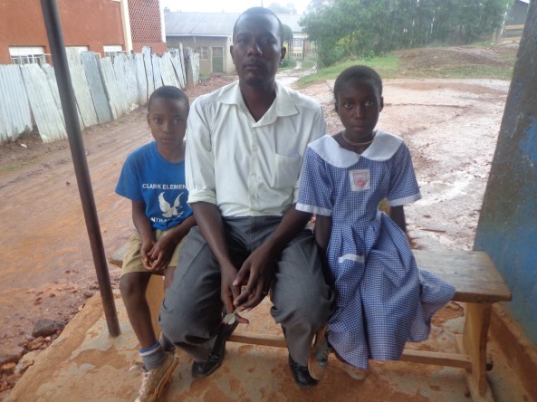 Richard Byamugisha, a CMM Team member and a school teacher at Sam Iga Primary School, sits with Kimera Wilson (boy on left side of picture) and Nanyange Cissy (girl on right side of picture). 